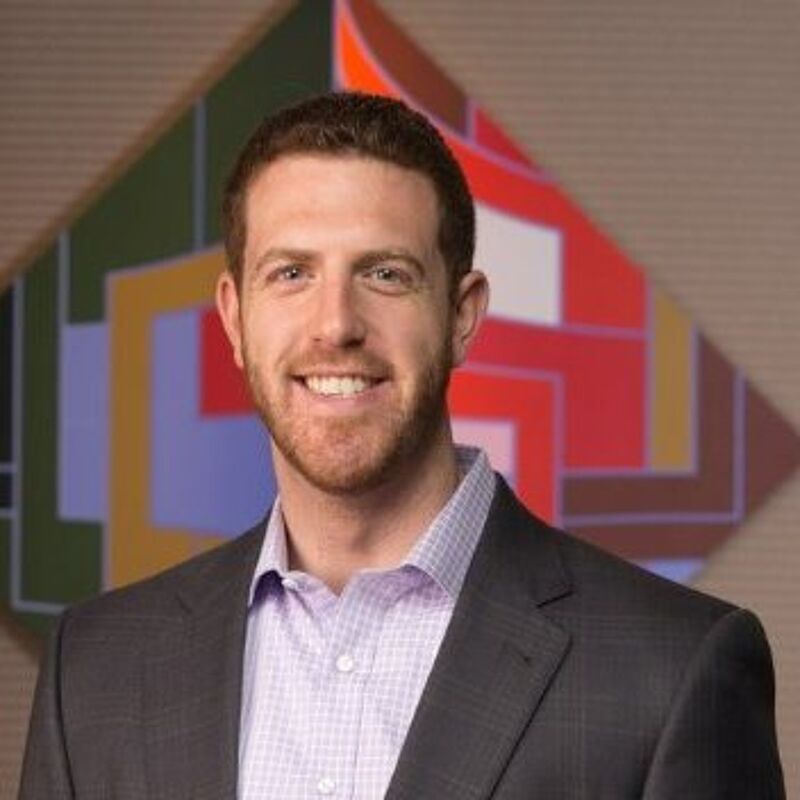 The image shows a man with short brown hair and a beard, wearing a suit jacket over a light purple shirt. He is smiling at the camera. Behind him is a colorful abstract painting. The background is a neutral color.